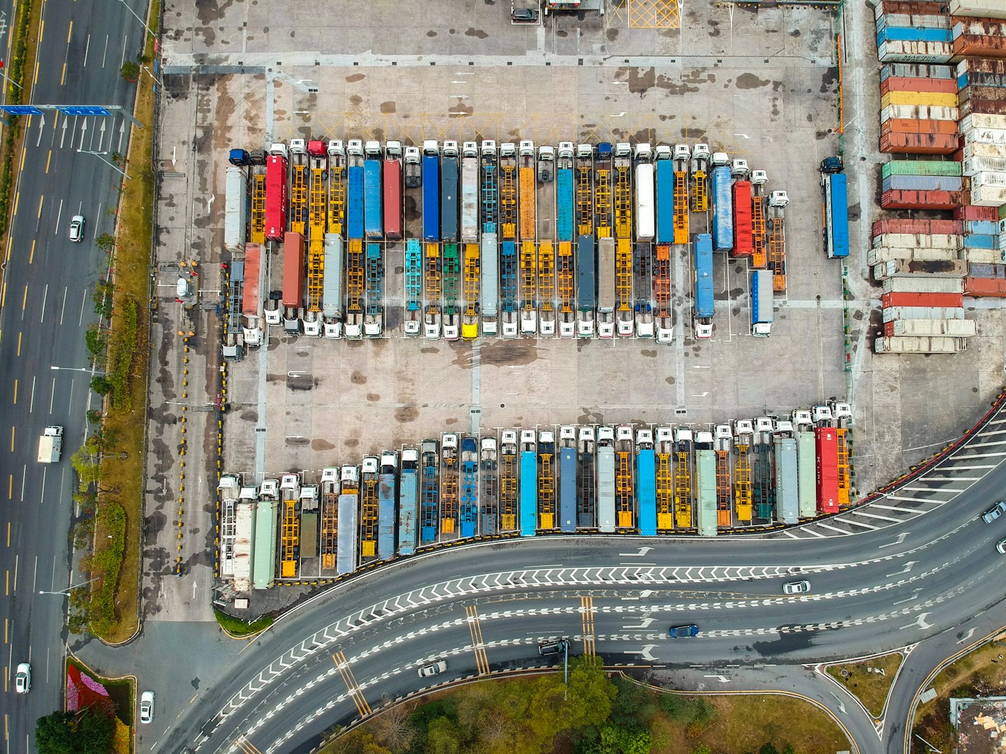 Na dużym placu logistycznym znajduje się wiele ciężarówek i ułożonych kontenerów.